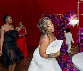 Smiling bride in white gown with fan, playful pose; guests and vibrant florals set a joyful wedding scene.