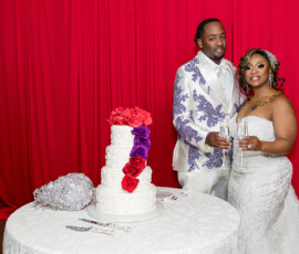Bride and groom in formalwear toast with champagne beside a floral wedding cake, bright red curtain behind them.