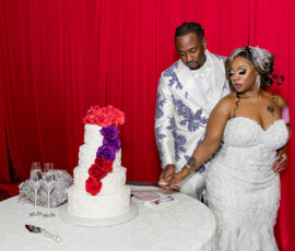 Bride and groom in formal attire cut a white cake with red and purple flowers beside a red curtain at their New Orleans wedding.
