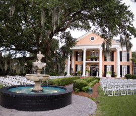 Peach mansion with white columns, white wedding chairs, and a stone fountain beneath an oak with moss—classic Louisiana style.