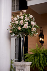 Glass vase with light pink roses, white flowers, and greenery on pedestal by tan brick house—ideal for wedding decor.