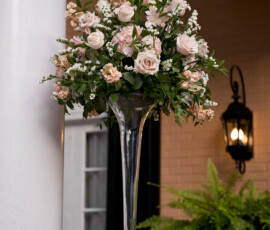 Glass vase with light pink roses, white flowers, and greenery on pedestal by tan brick house—ideal for wedding decor.