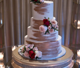 Elegant three-tiered white cake with rose gold brush strokes, red and pink roses, and votive candles on a mirrored table.