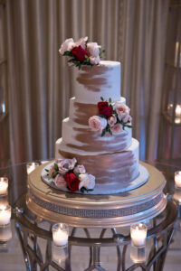 Elegant three-tiered white cake with rose gold brush strokes, red and pink roses, and votive candles on a mirrored table.