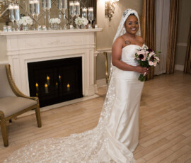 Bride in white gown and floral veil smiles with bouquet in elegant New Orleans room with chandelier, fireplace, and mirrors.
