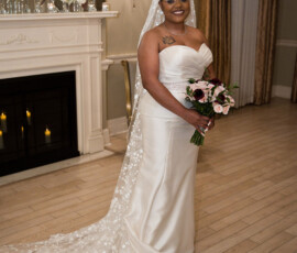 Bride in white gown with lace veil holds bouquet, smiling by a decorated fireplace at an elegant Gulf South wedding.