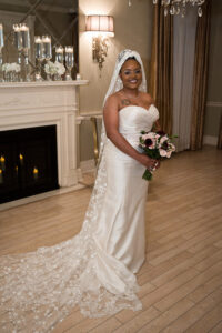 Bride in white gown with lace veil holds bouquet, smiling by a decorated fireplace at an elegant Gulf South wedding.