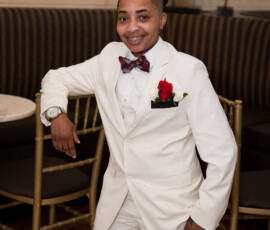 Smiling person in a white suit with red rose boutonniere, leaning on a gold chair at an elegant, candlelit wedding reception.