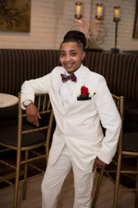 Smiling person in a white suit with red rose boutonniere, leaning on a gold chair at an elegant, candlelit wedding reception.