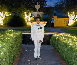 Person in a white suit walks a light-lined path with hedges, fountain, and trees at an elegant Louisiana wedding reception.