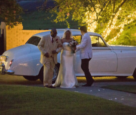 Bride in white dress with two in white suits by classic car at dusk, tree lights glowing—Gulf South wedding inspiration.