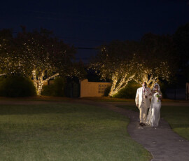 Bride and older man walk a curved, tree-lit path at night, grass on both sides, creating a magical Gulf South wedding vibe.