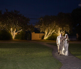 Bride in white dress and man in suit walk at night on a lit path, surrounded by string-lit trees at a Louisiana wedding.