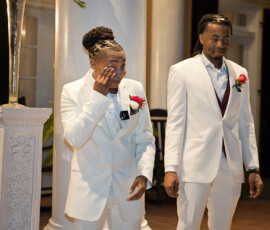 Two people in white suits with red rose boutonnieres stand by a pillar at a touching New Orleans-inspired wedding ceremony.
