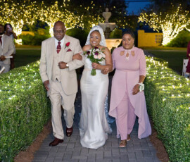 A bride in a white dress walks an outdoor aisle at night, holding a bouquet, with two people and string lights in Louisiana.
