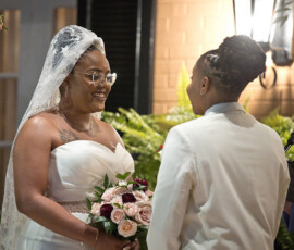 Two women smile at each other during a Louisiana wedding, one in a white dress with roses, the other in a white suit jacket.