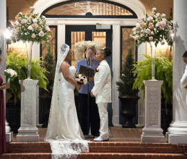 Two women marry at an outdoor altar, one in a gown and veil, the other in a suit, with attendants and floral decor.