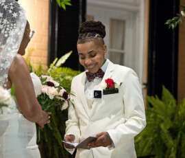 A happy couple stands at their wedding ceremony, one in a white suit reading from a card, the other in a lace veil and gown.