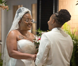 Two women exchange vows outdoors in New Orleans, smiling and holding hands in wedding attire amid greenery and warm light.