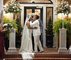A couple in white suits shares a kiss at their New Orleans-inspired wedding with floral decor and attendants nearby.