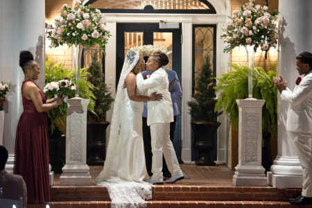 A couple in white suits shares a kiss at their New Orleans-inspired wedding with floral decor and attendants nearby.