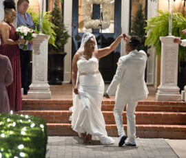 Newlyweds in white dance outside at their Louisiana wedding, smiling as guests and bridesmaids watch amidst evening greenery.