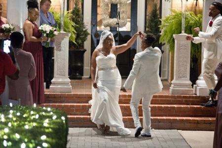 Newlyweds in white dance outside at their Louisiana wedding, smiling as guests and bridesmaids watch amidst evening greenery.