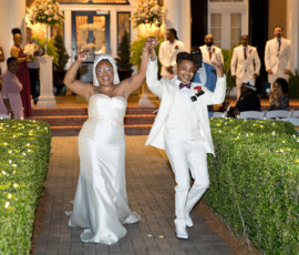 A joyful couple in white walks hand in hand down a nighttime outdoor aisle as guests smile, celebrating their Louisiana wedding.