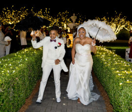 Newlyweds in white walk under twinkling lights with umbrellas, cheered by guests—a magical Louisiana-inspired wedding moment.