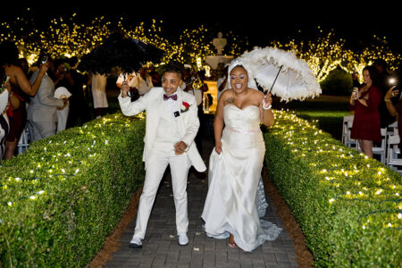 Newlyweds in white walk under twinkling lights with umbrellas, cheered by guests—a magical Louisiana-inspired wedding moment.