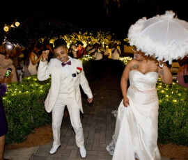 Newlyweds in formal wear dance with parasols outdoors at night, surrounded by guests and string lights at a Gulf South wedding.