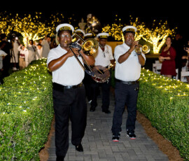 A brass band in white shirts and hats marches at night along a hedge-lined, fairy-lit path during a Gulf South wedding.