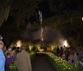 Guests at an outdoor event watch fireworks near a garden fountain, with glowing lights and trees framing this magical New Orleans scene.