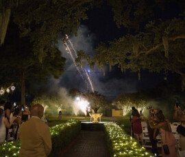 Guests at an outdoor evening wedding watch fireworks near a lit fountain, surrounded by trees and glowing string lights.