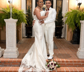 Two brides in white embrace and smile on brick steps, with floral decor and bouquet, celebrating their wedding day.