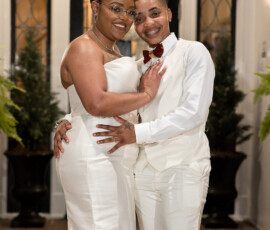 Smiling couple in white wedding attire embraces by a plant-adorned doorway, perfect for celebration inspiration.