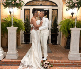 Two brides in white embrace on brick steps with floral decor, smiling and capturing Gulf South wedding inspiration.