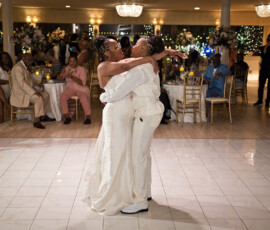 Two brides in white dance and smile on a ballroom floor as guests watch and take photos at a New Orleans wedding.
