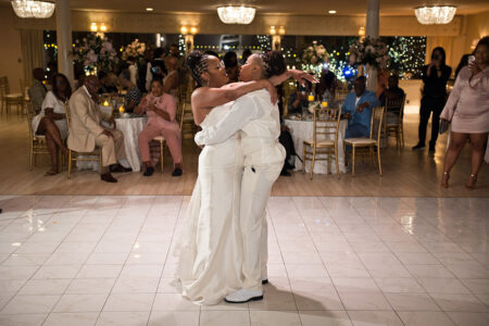 Two brides in white dance and smile on a ballroom floor as guests watch and take photos at a New Orleans wedding.