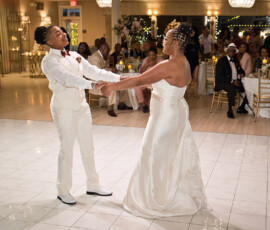 Two brides dance, smiling and holding hands at their elegant Gulf South wedding reception, surrounded by guests.