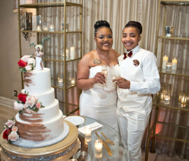 Smiling couple in white hold champagne by a three-tiered floral wedding cake, elegant New Orleans celebration with candles.