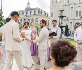 Bride and groom hold hands at an outdoor Louisiana wedding with guests, wedding party, and historic buildings in the background.
