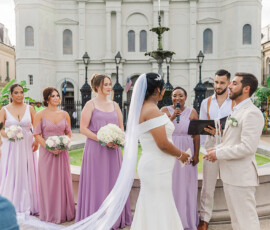 Bride and groom exchange vows outdoors with lavender bridesmaids, light-suited groomsmen, fountain, and historic building behind.