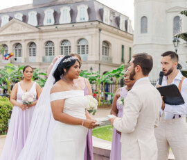 Bride and groom exchange vows outdoors with bridesmaids in lavender, officiant, historic building, and lush greenery.