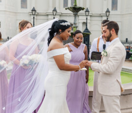 Bride in white and groom in beige exchange rings at outdoor Louisiana wedding, bridesmaids in lavender nearby.