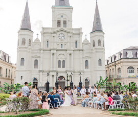 Outdoor wedding at a historic Louisiana cathedral with three spires, gardens, and guests under a cloudy sky.