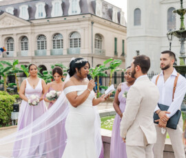 Bride in white reads vows to groom at outdoor Gulf South wedding, with lavender bridesmaids and historic building backdrop.
