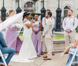 Bride in white reads vows to groom at elegant New Orleans outdoor wedding, with lavender bridesmaids and beige groomsmen by a fountain.