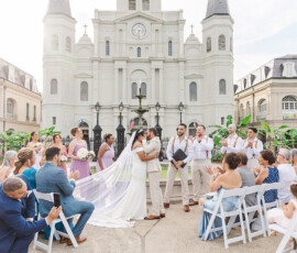 A couple kisses at their outdoor New Orleans church wedding, surrounded by applauding guests and their wedding party.