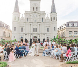 Bride and groom walk down an outdoor aisle by a historic cathedral, smiling at guests—romantic Louisiana wedding inspiration.